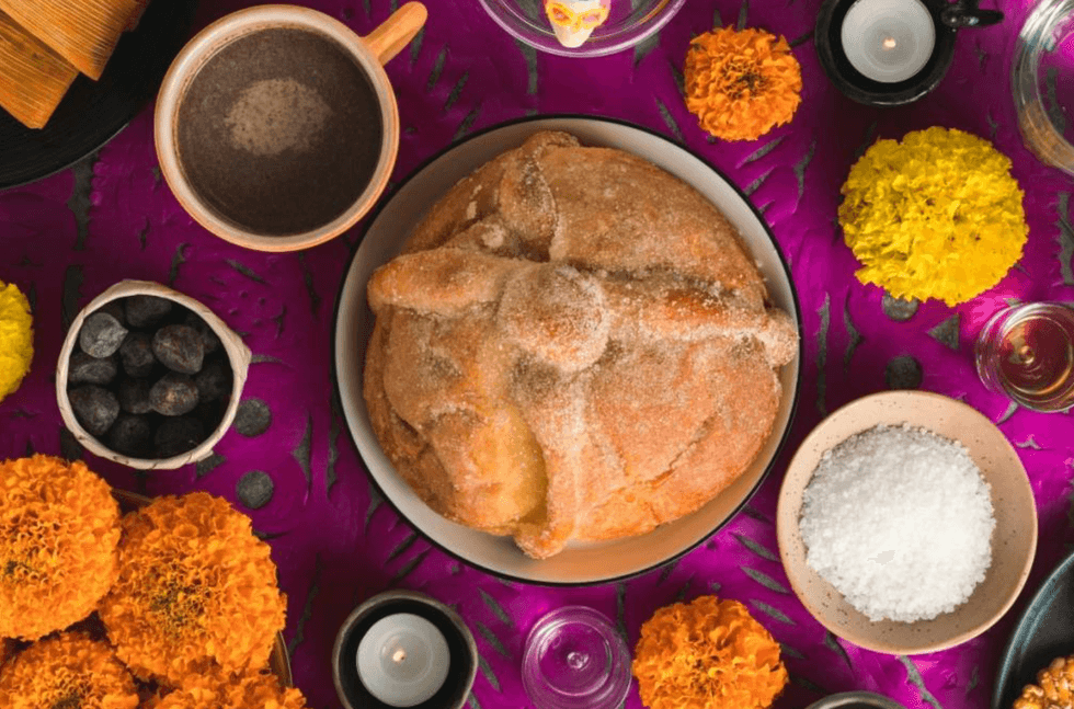 Pan de muerto, candles, marigolds, chocolate, and seeds on a purple tablecloth.