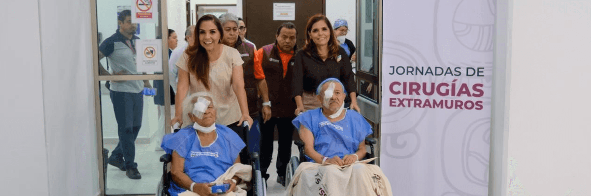 Patients in wheelchairs exit surgery area with staff assistance, near medical event banner.