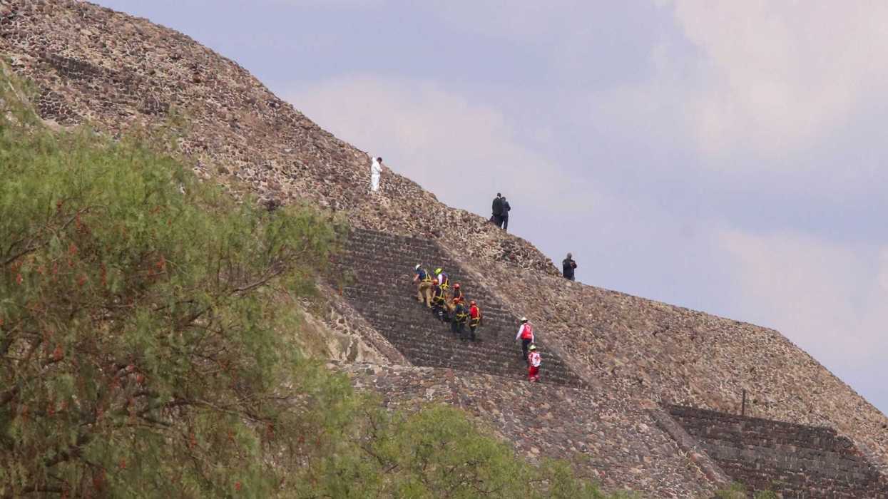 People climbing the steep steps of an ancient stone pyramid under a blue sky.