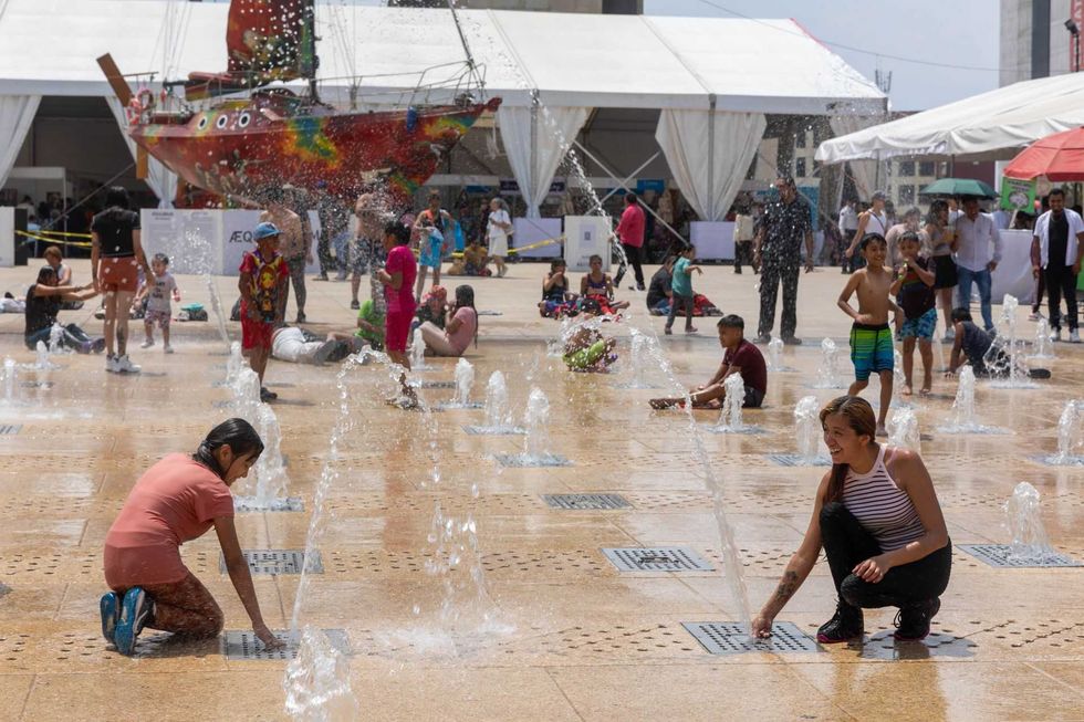 People enjoying a fountain at a public square with a colorful boat in the background.