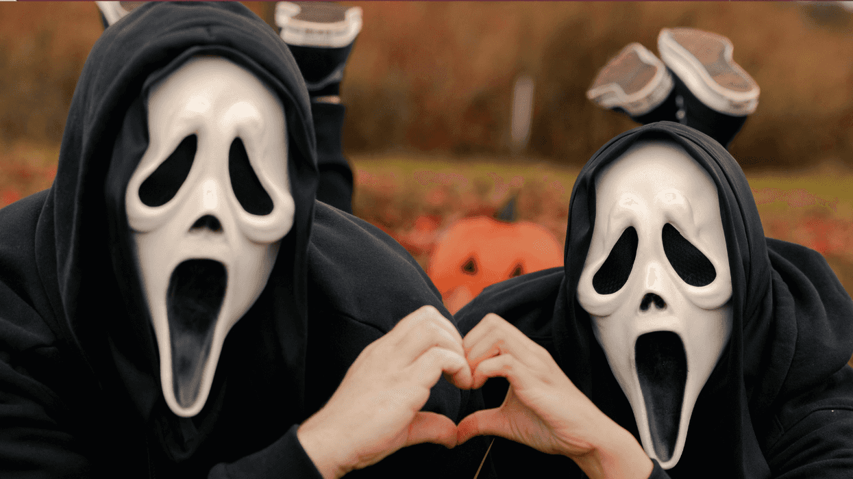 People in Scream masks making a heart with their hands, laying on ground with pumpkins behind.