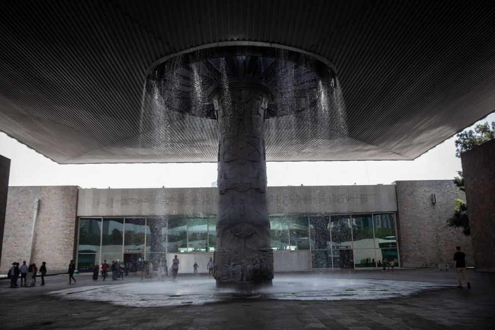 People under large metal roof with central column waterfall in courtyard.