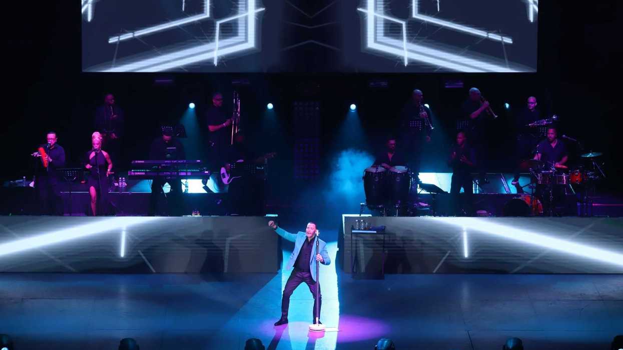 Performer sings on stage with live band and geometric light display above.