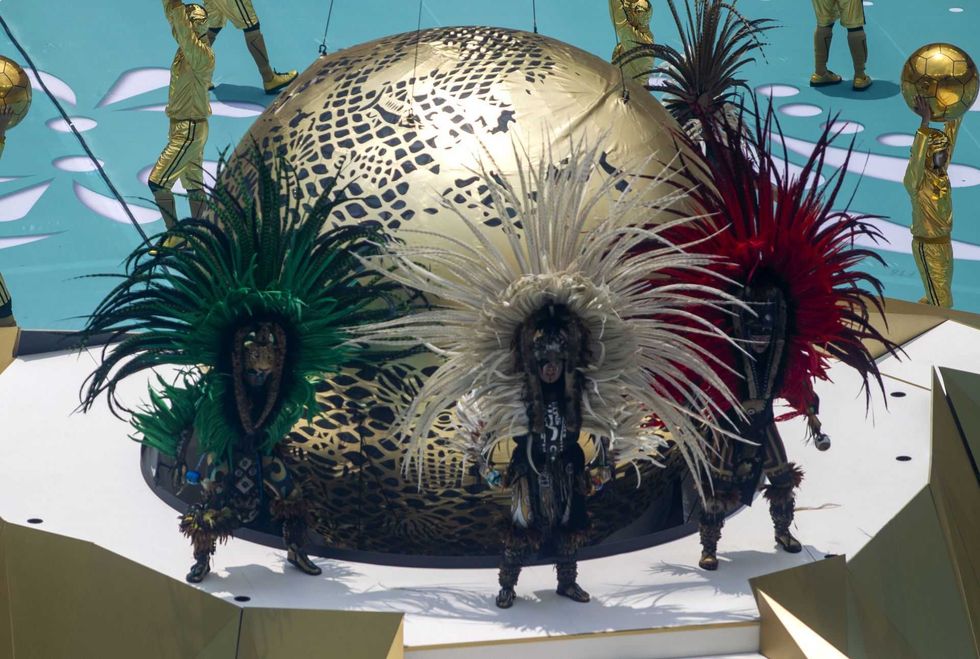 Performers in vibrant feathered headdresses stand on stage with a golden sphere backdrop.