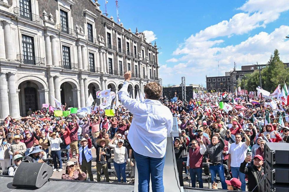 Person addressing large crowd at an outdoor rally beside a historic building.