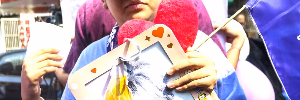 Person holding a framed photo and a purple flag at a public event, wearing a purple cap.