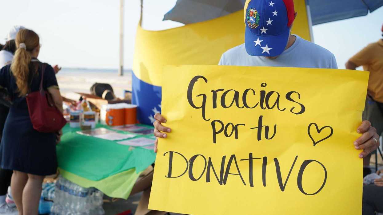 Person holds yellow "Gracias por tu donativo" sign at donation stand with Venezuelan flag.