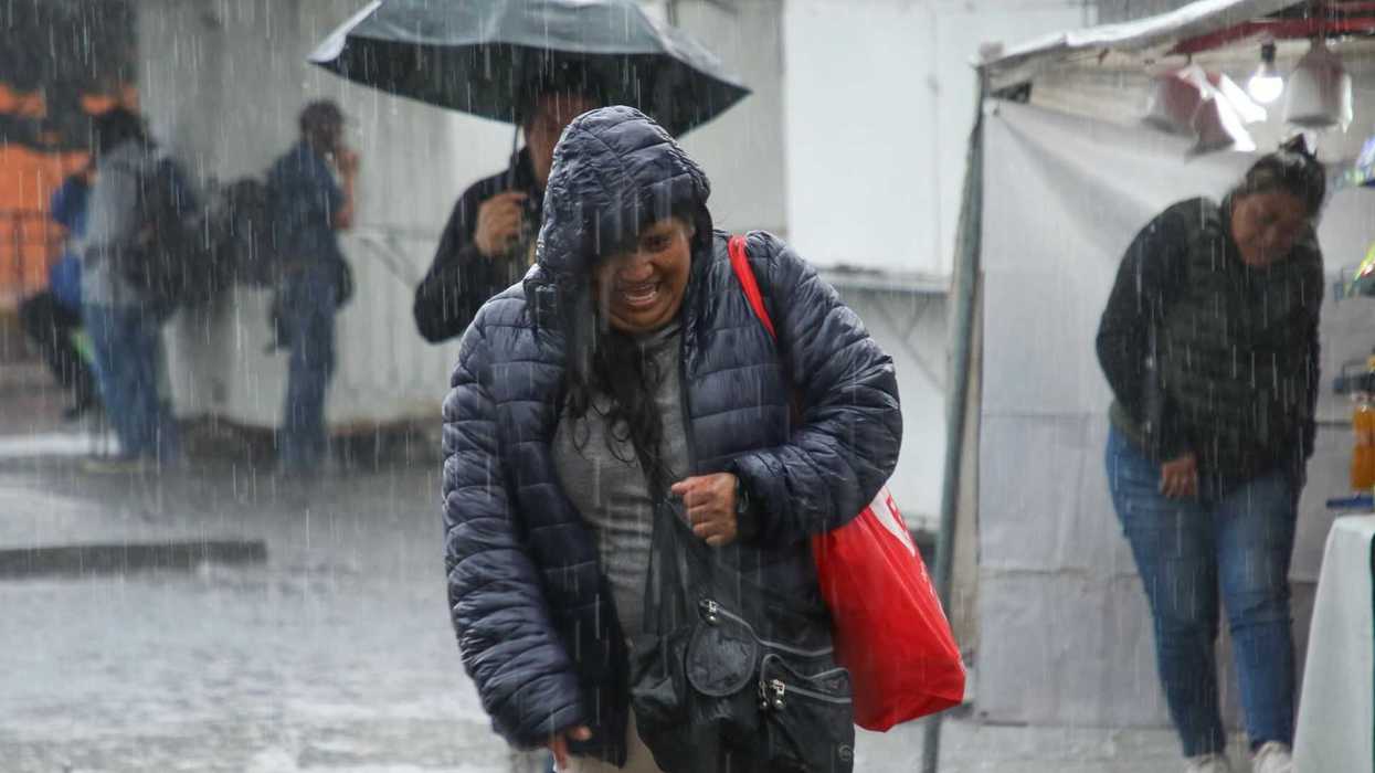 Person in a blue jacket struggles through rain with a red bag, while others take cover.