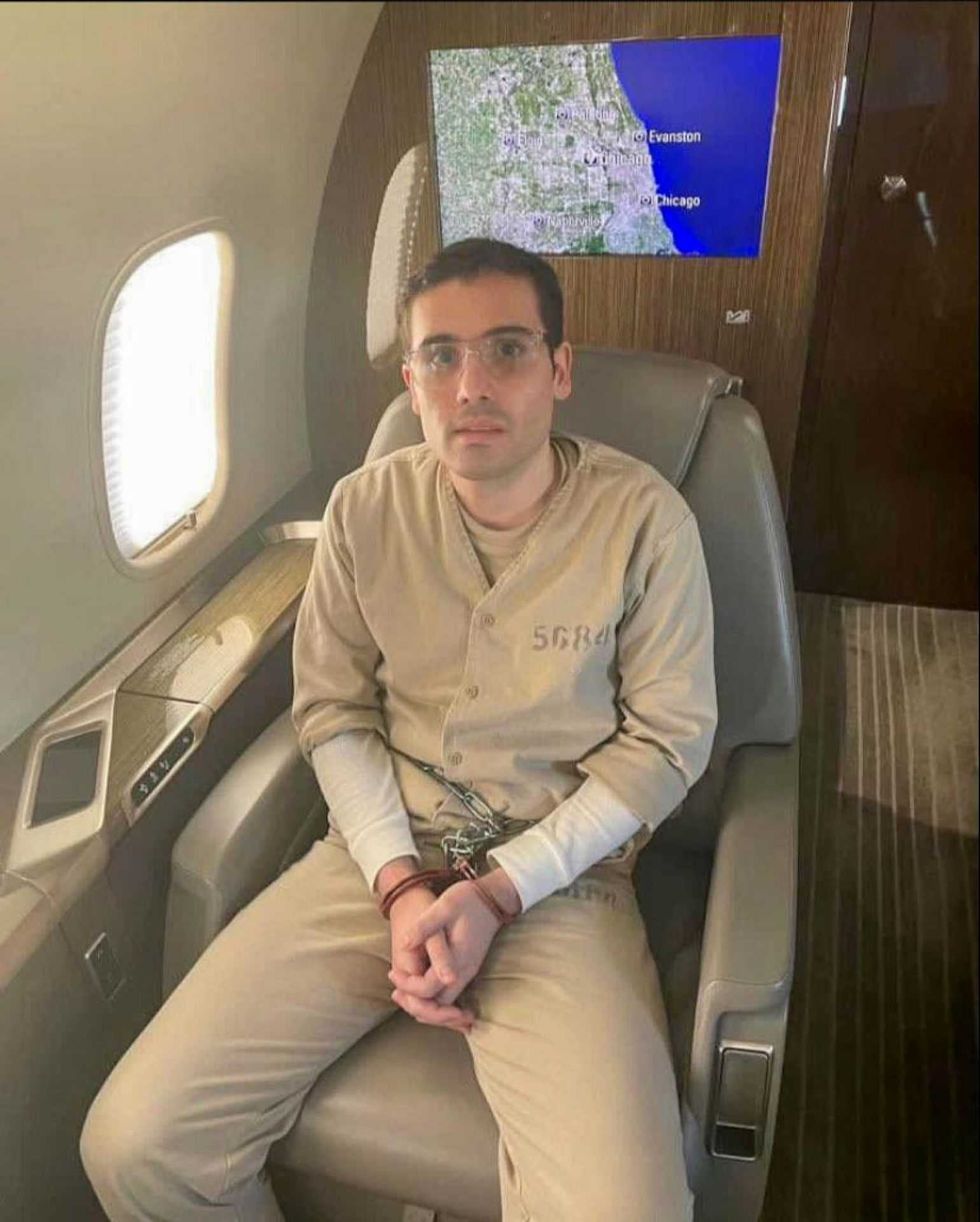 Person in beige prison jumpsuit sitting on plane in handcuffs, map visible in background.