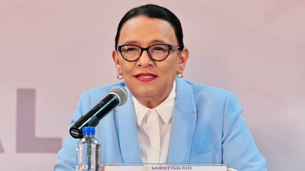 Person in blue suit speaking at a microphone, with a backdrop and water bottle.