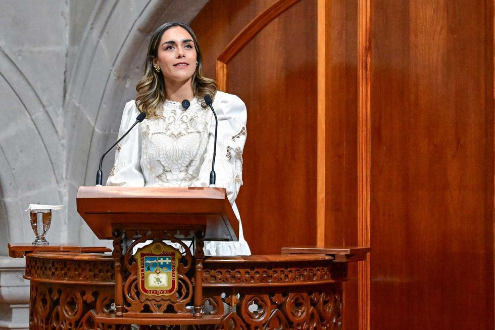 Person in white dress speaking at ornate podium in a formal setting.
