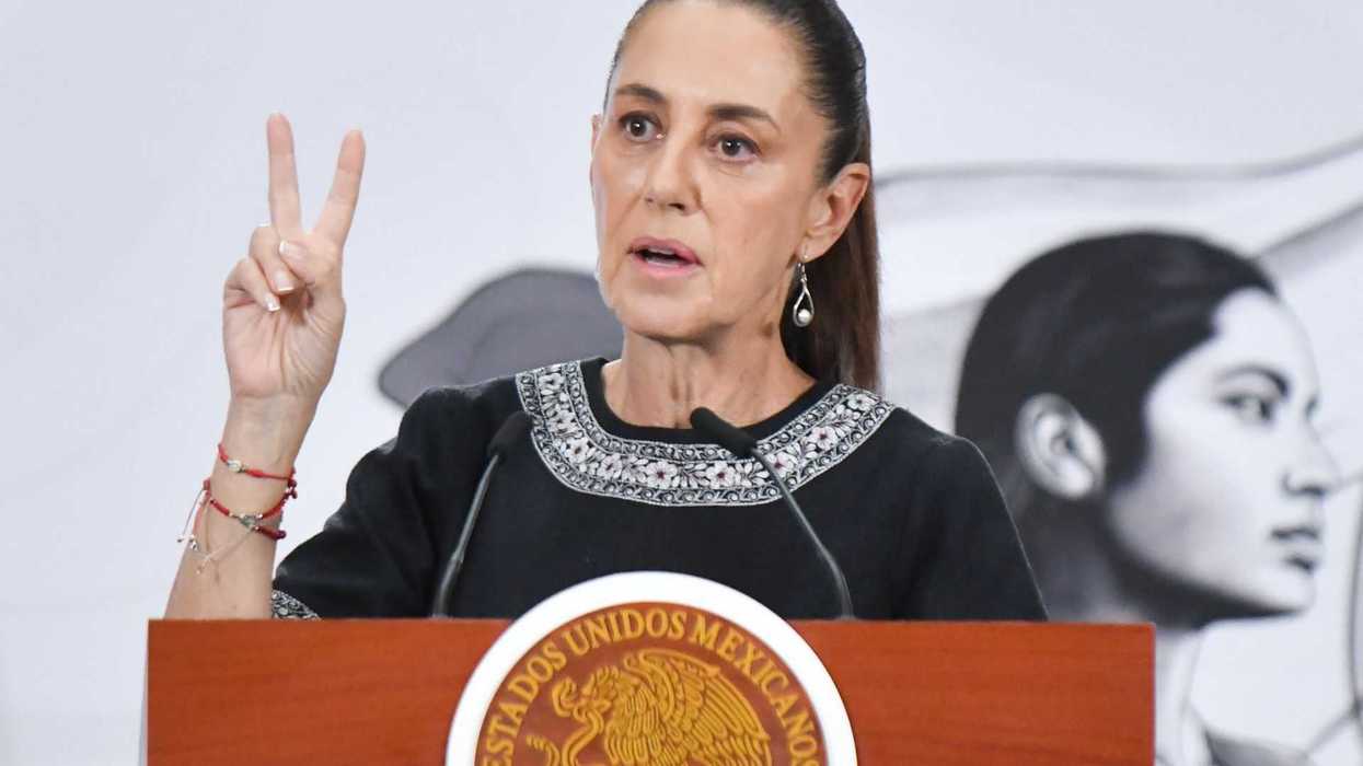 Person speaking at a podium, gesturing peace sign, with a mural in the background.