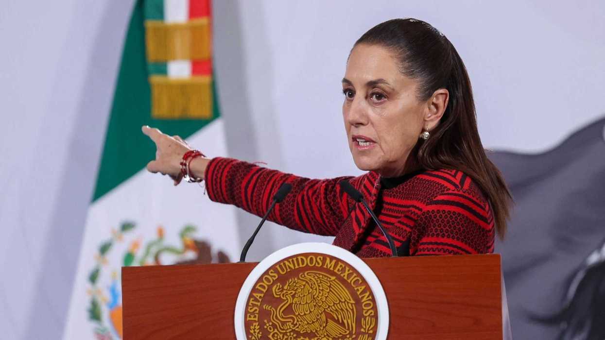 Person speaking at a podium, gesturing, with a Mexican flag in the background.