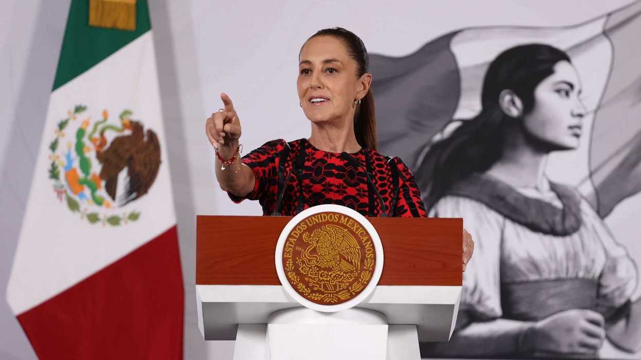 Person speaking at a podium with Mexican flag and mural in background.