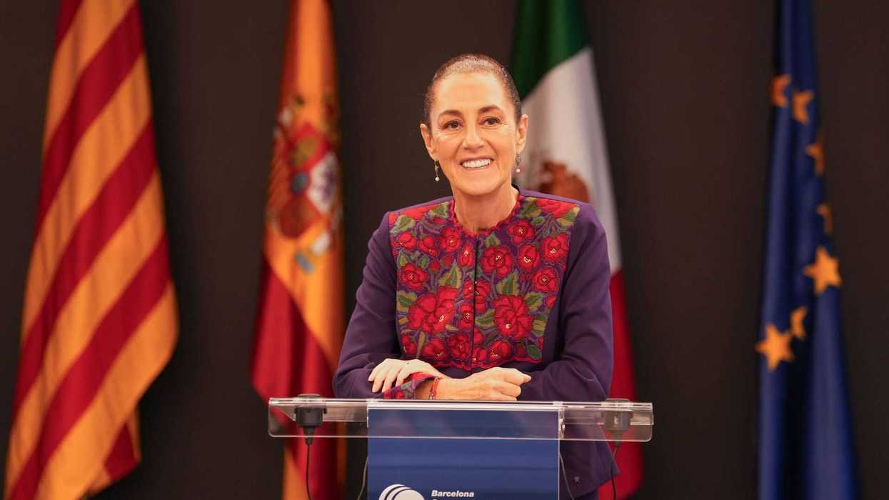 Person speaking at podium with floral attire, flags in the background.