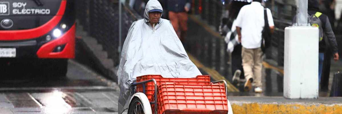 Person with poncho riding tricycle in rainy city street near bus and pedestrians with umbrellas.