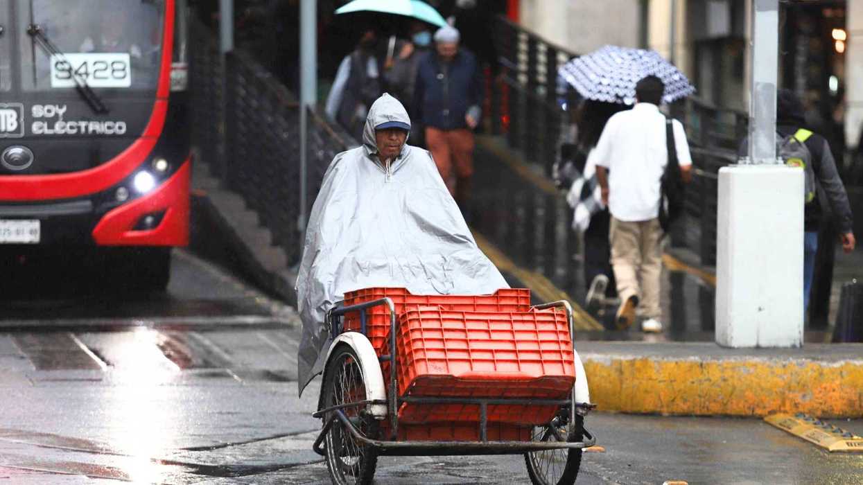 Person with poncho riding tricycle in rainy city street near bus and pedestrians with umbrellas.