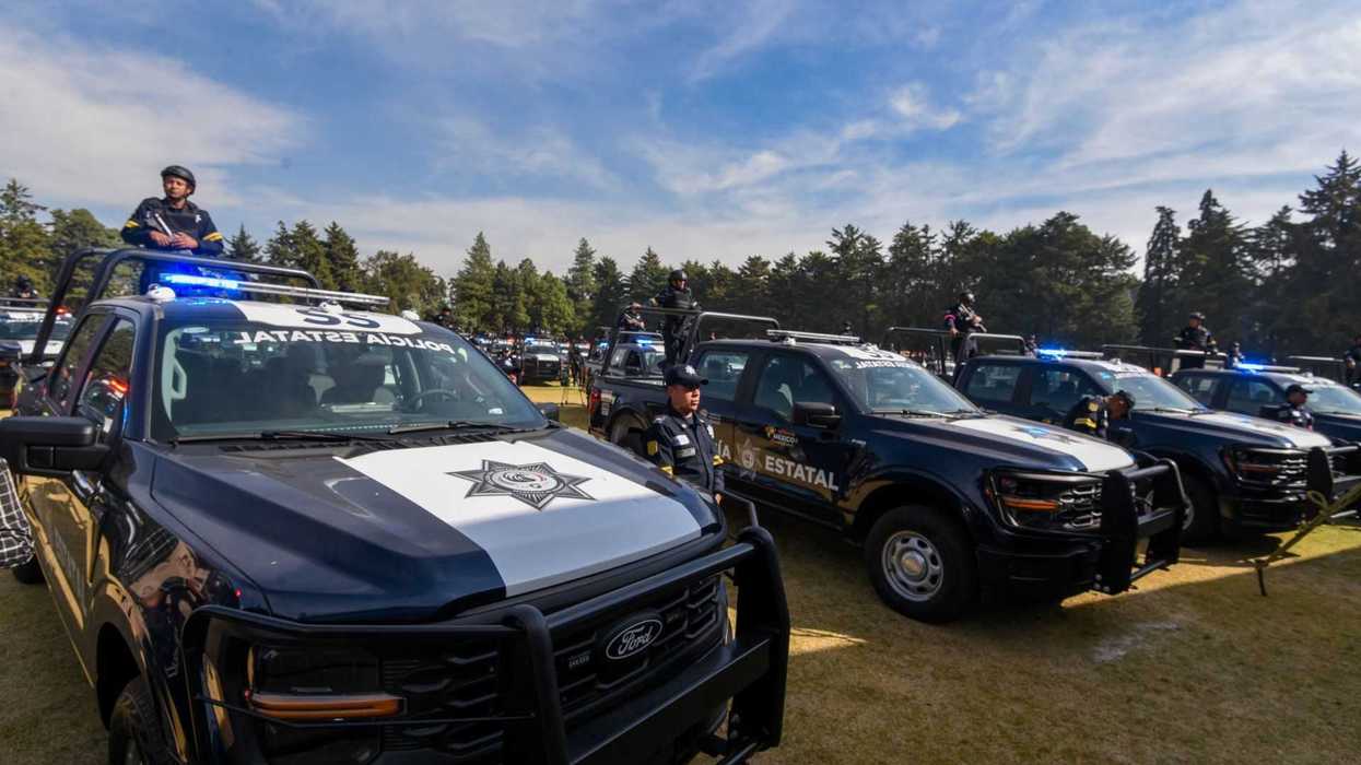 Police vehicles lined up with officers standing nearby in a grassy area under a blue sky.