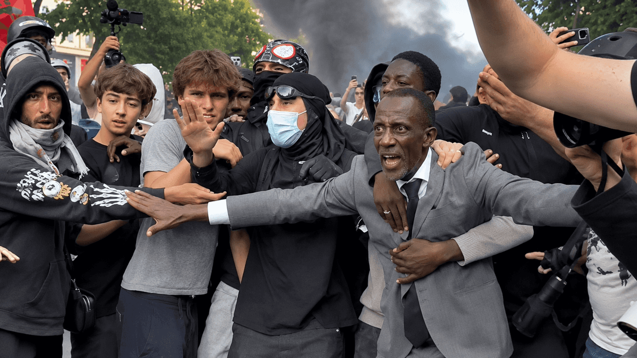 Protesters and a man in a suit in a tense standoff, smoke rising in the background.