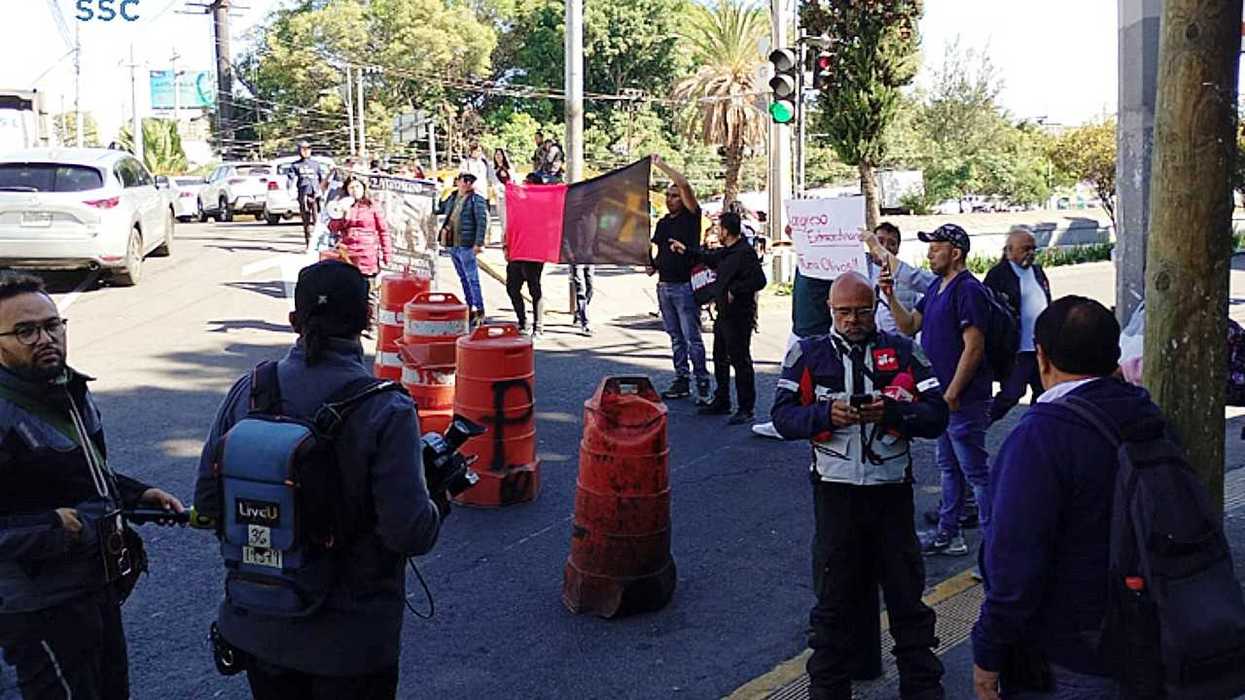Protesters block street with signs and traffic cones, people and cars in the background.