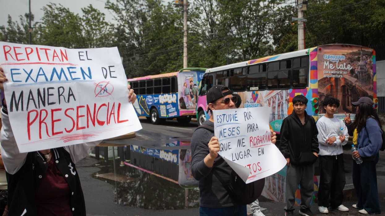 Protesters hold signs demanding in-person exams, with colorful buses in the background.
