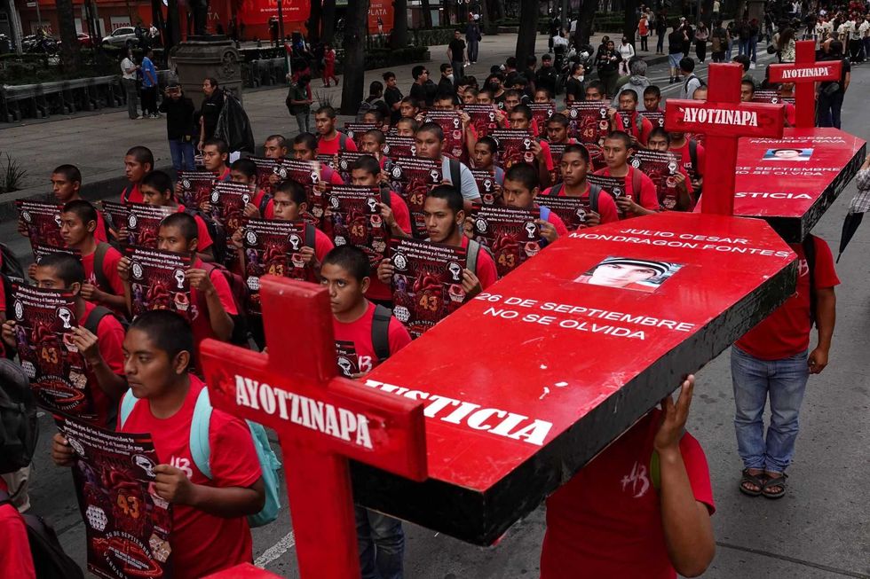 Protesters in red shirts hold signs and large red crosses, commemorating Ayotzinapa victims.