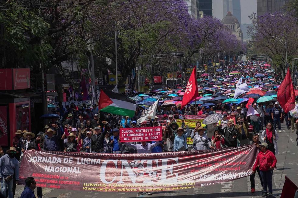 Protesters march down a city street with banners and flags under colorful umbrellas.