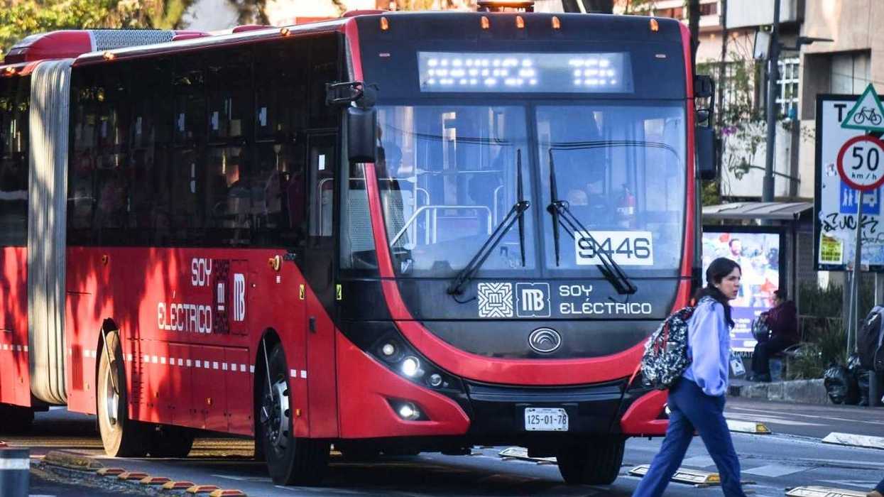 Red electric bus on city street, woman crossing, speed limit signs visible.