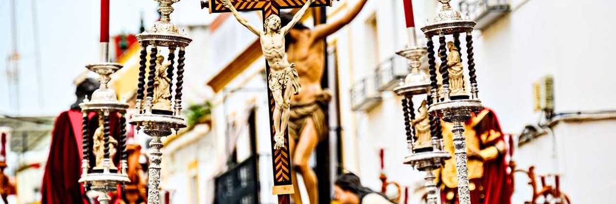 Religious parade with crucifix and ornate candle holders in a sunny street.