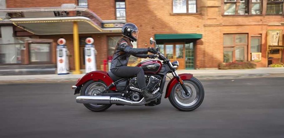 Rider on a red motorcycle passing by vintage gas pumps and brick buildings.