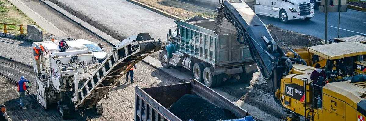 Road construction with heavy machinery and trucks, traffic in the background.