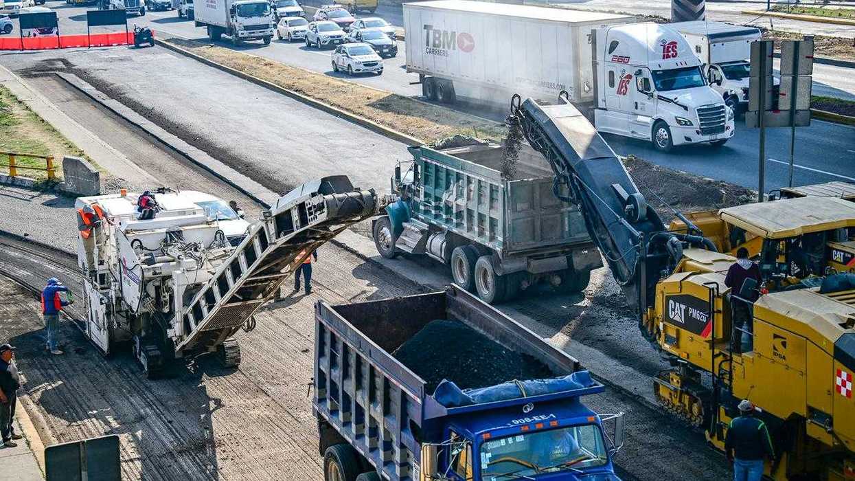 Road construction with heavy machinery and trucks, traffic in the background.