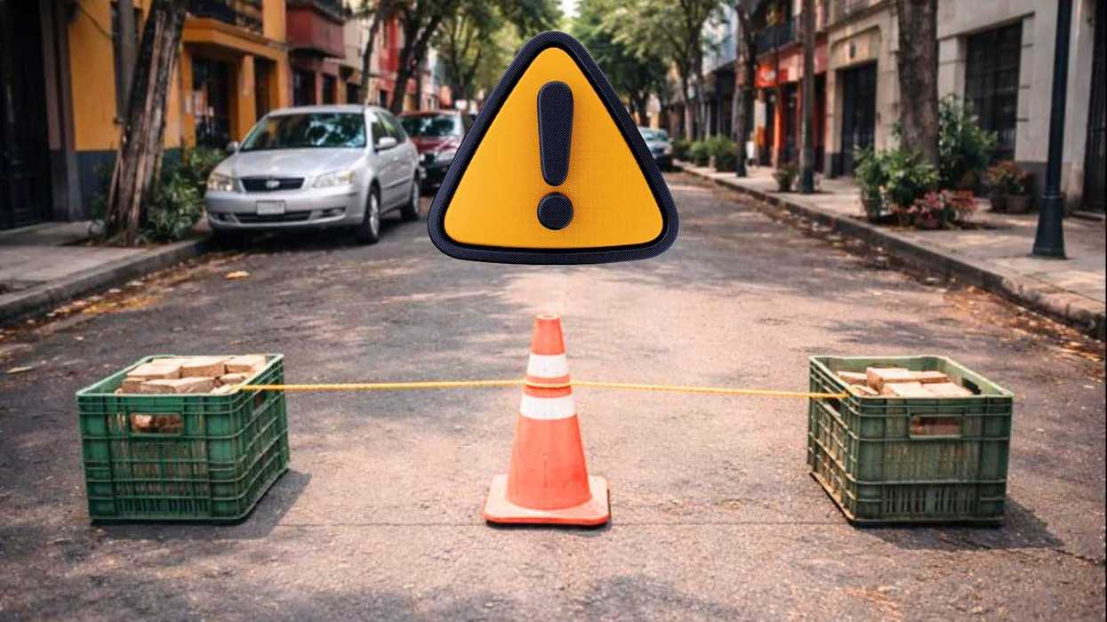 Roadblock with cone and crates on urban street, warning sign above.