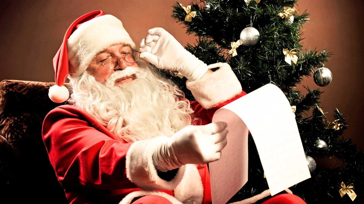 Santa reading a long list beside a decorated Christmas tree.