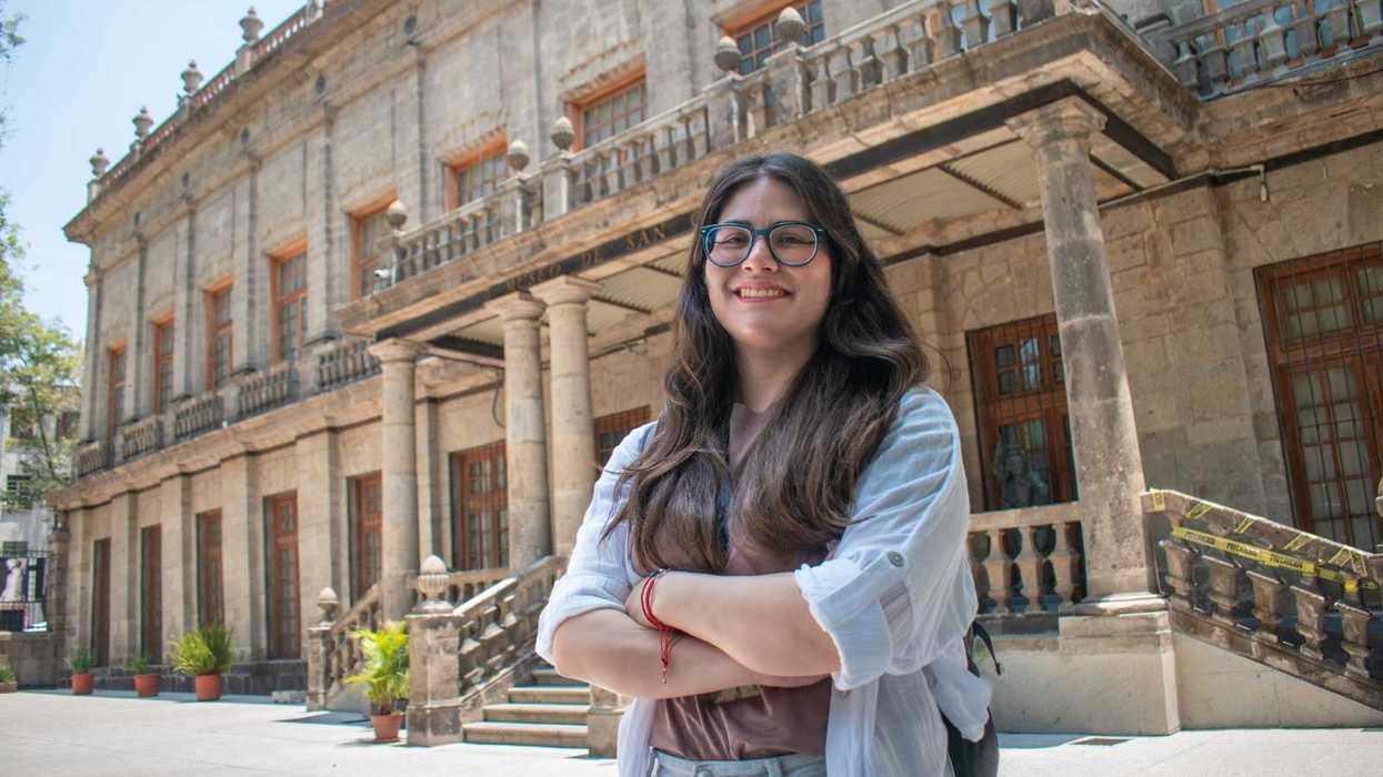 Smiling person with long hair stands before a historic stone building with balconies.