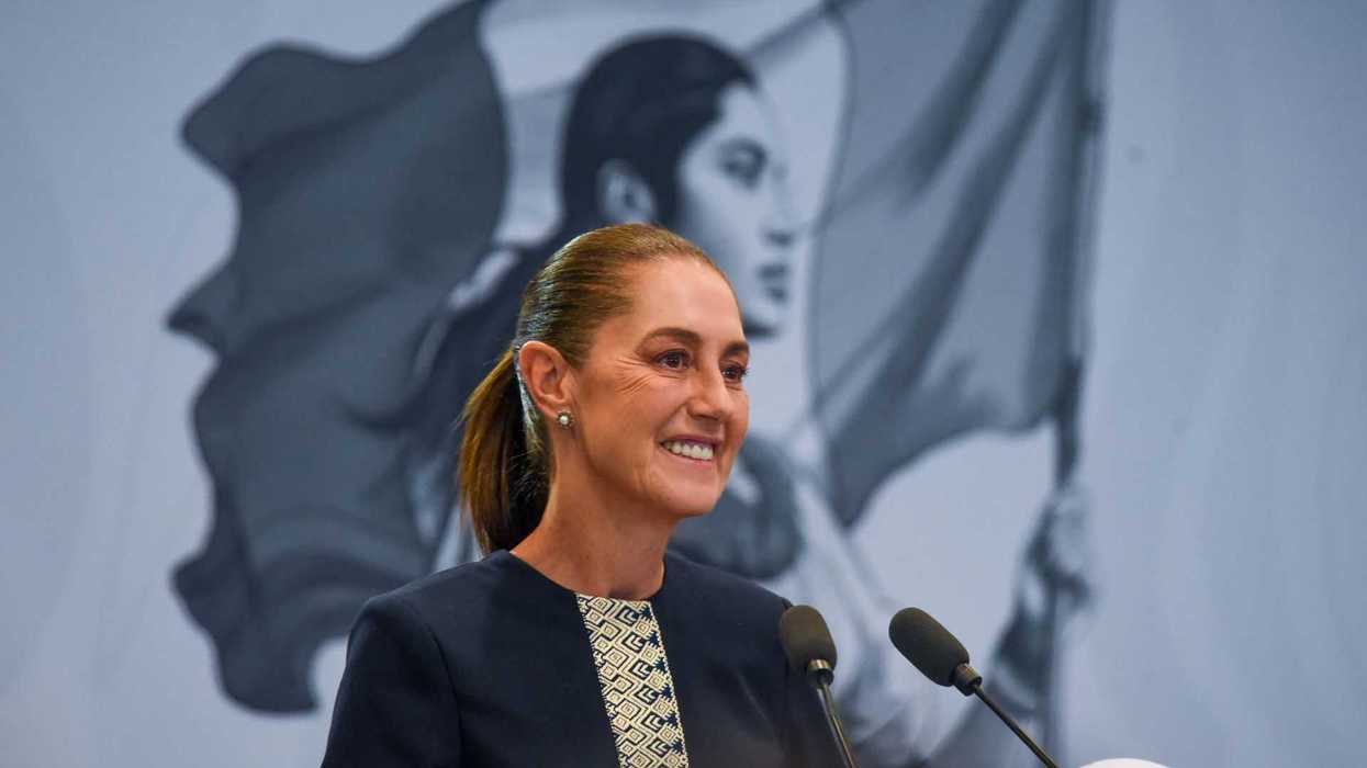 Smiling woman at podium, Mexican flag mural in background.