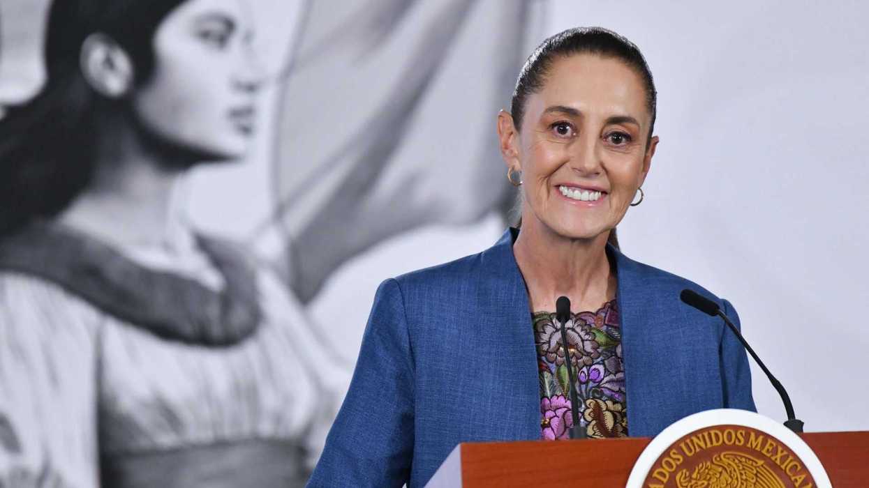 Smiling woman in blue jacket speaking at podium, historical female portrait in background.