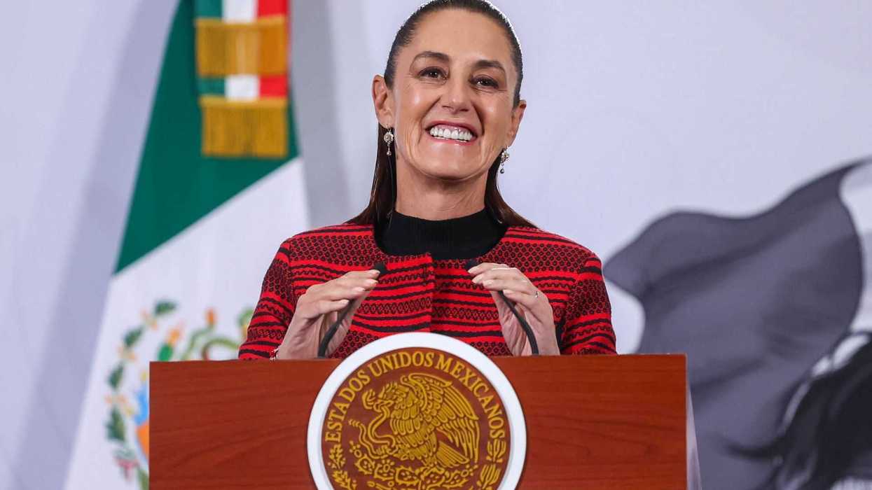 Smiling woman speaks at podium with Mexican flag in the background.