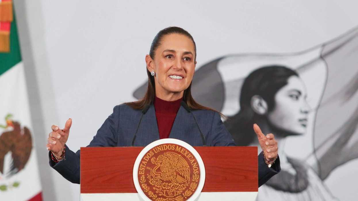 Smiling woman speaks at podium with Mexico's emblem, flag background.