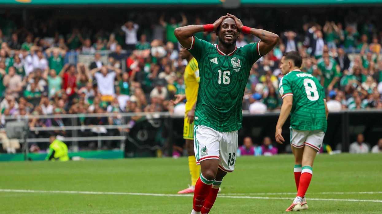 Soccer player in green celebrates on field, hands on head, crowd in background.