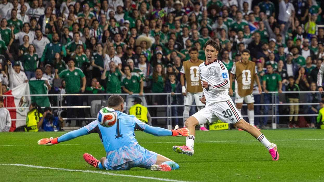 Soccer player in white about to score against a diving goalie in light blue, with cheering crowd.