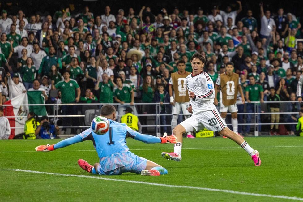 Soccer player in white about to score against a diving goalie in light blue, with cheering crowd.