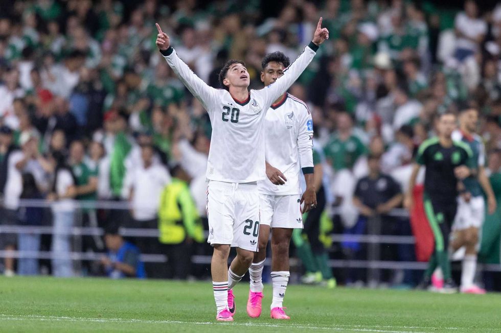 Soccer player in white kit celebrates with arms raised; crowd in background.