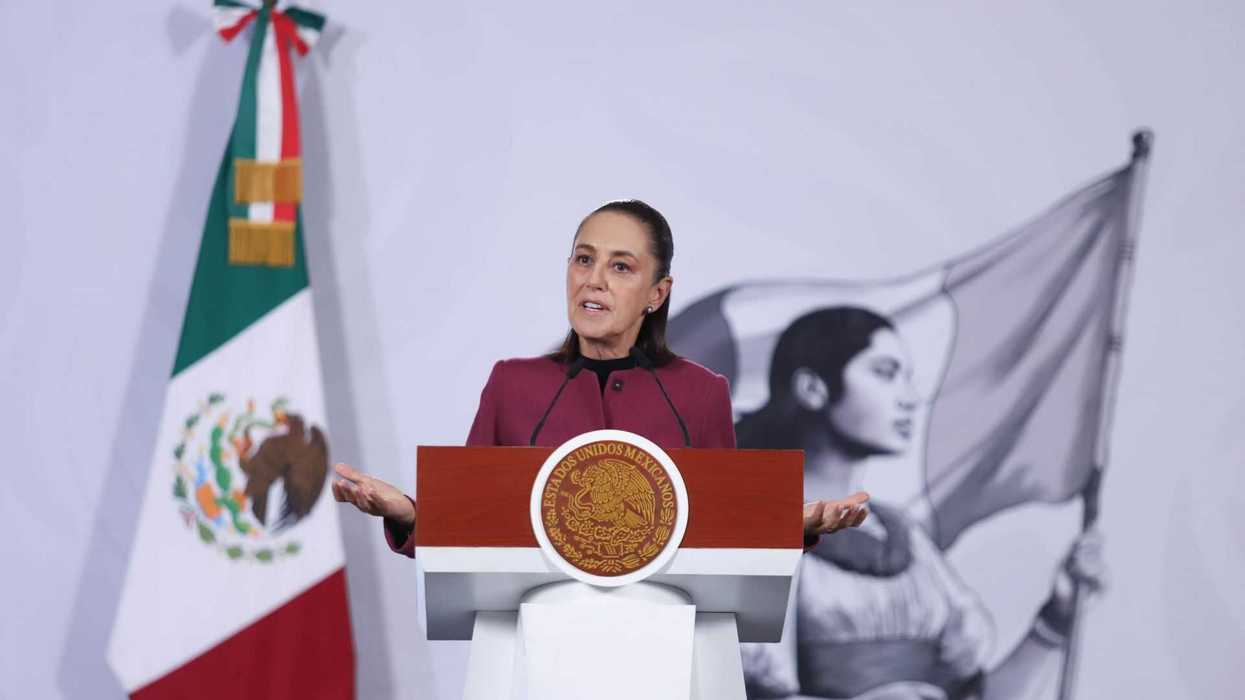 Speaker at podium with Mexican flag, mural of woman holding flag in background.