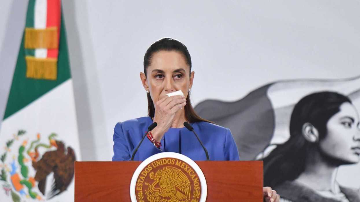 Speaker in blue at podium with Mexican seal, flag and mural in background.