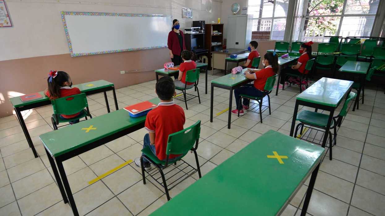 Students in masks sit apart in a classroom with distanced desks and a masked teacher.