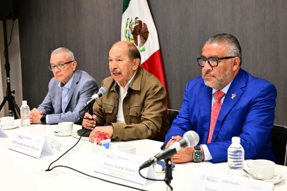 Three men at a meeting table with microphones, Mexican flag in background.