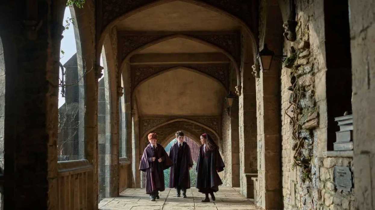 Three people in cloaks walk through a medieval stone corridor with arches.