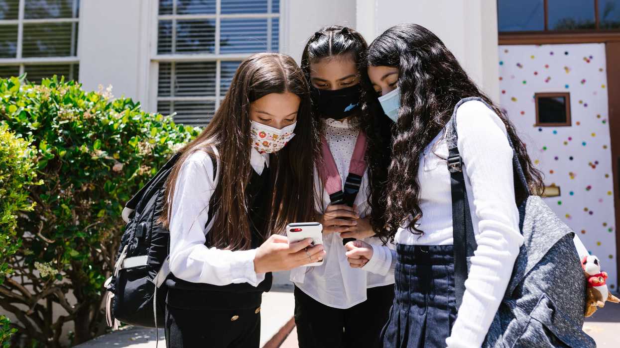 Three young students in masks look at a phone outside a school building.