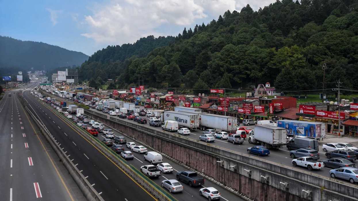 Traffic jam on highway near shops and forested hills under a cloudy sky.